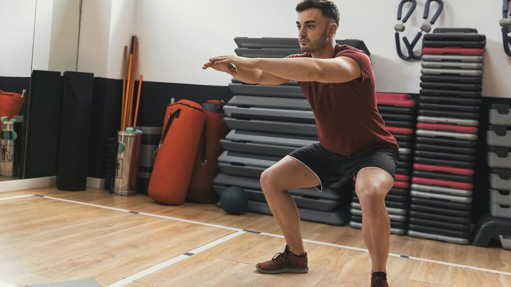 Man doing a bodyweight squat with arms extended in a bright gym studio, a beginner-friendly exercise