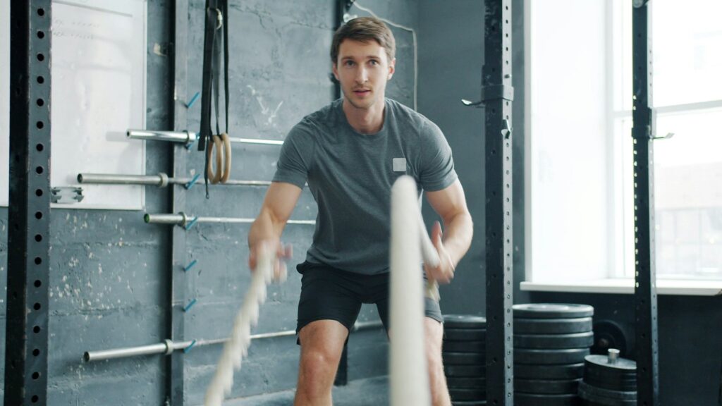 Man training with battle ropes in a functional gym with racks, gymnastic rings and weight plates
