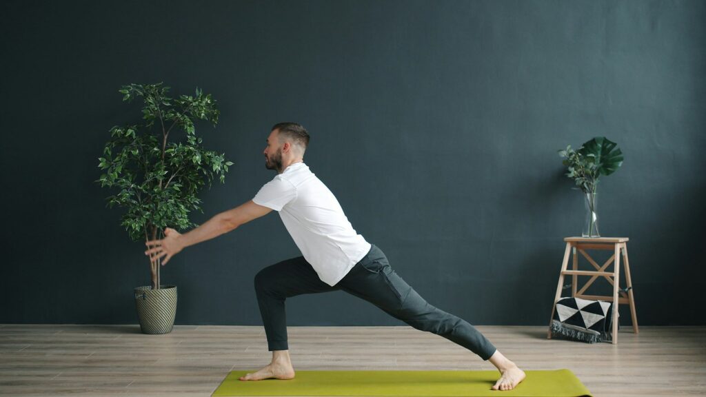 Man in a t-shirt and track pants doing a deep lunge stretch on a mat for mobility and recovery